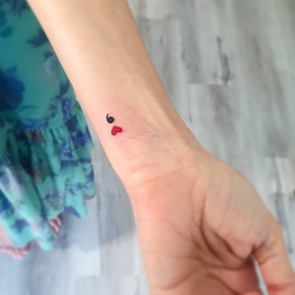 Semicolon tattoo on the wrist, replacing the dot with a heart shape filled in red, symbolizing resilience and love: heartfelt pause 